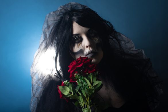 A woman with skull makeup and a black veil holds a bouquet of roses and looks at the camera gloomily