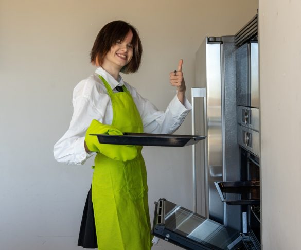 A smiling woman wearing an apron, putting a baking tray into an oven as she gives a thumbs up