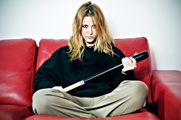 A blonde woman with an angry expression on her face, sitting cross-legged on a sofa. She is holding what looks like a baseball bat in both hands.