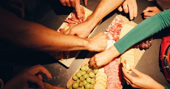 A top down view of a charcuterie board with cheese, grapes and meat, with lots of hands reaching across to take food