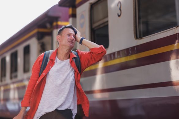 A man standing by a train wearing a backpack. He looks stressed and is holding his head with one hand
