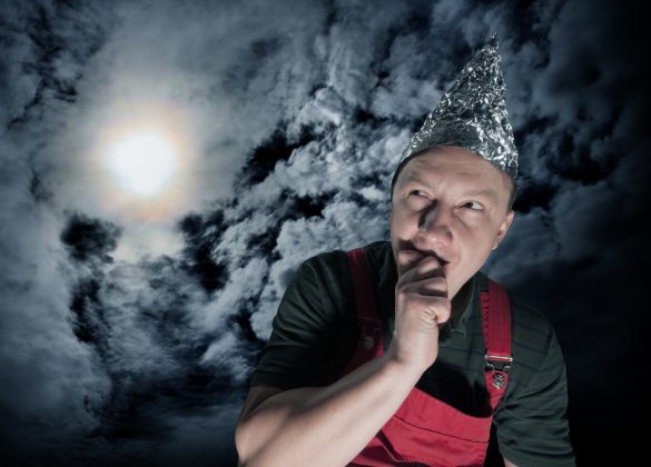 A man wearing a tin foil hat stands in front of a cloudy night sky, looking suspiciously at the moon
