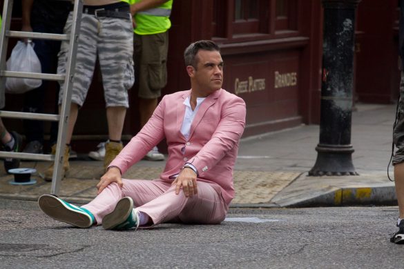 Singer Robbie Williams wearing a pink two-piece suit and sitting down by a street curb.
