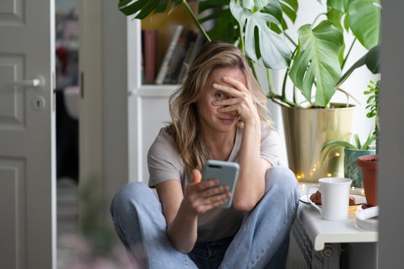 A woman sitting on the floor peering through her fingers as she looks at a phone.