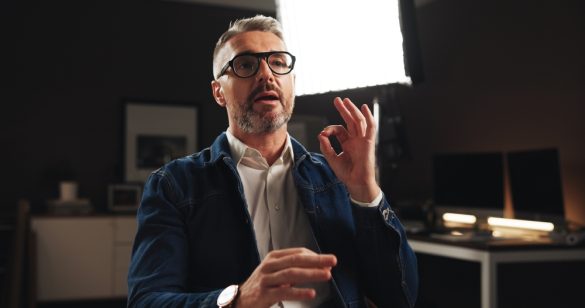 A man with short hair and glasses wearing a blazer. He is sitting oriented towards the camera and is in the middle of talking.
