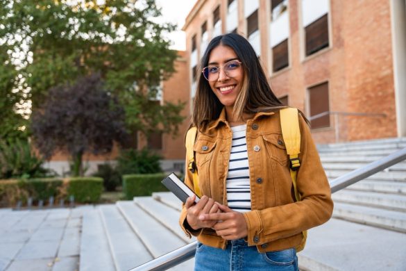 A woman wearing a backpack and standing on some outdoor steps, holding a book under one arm and smiling.