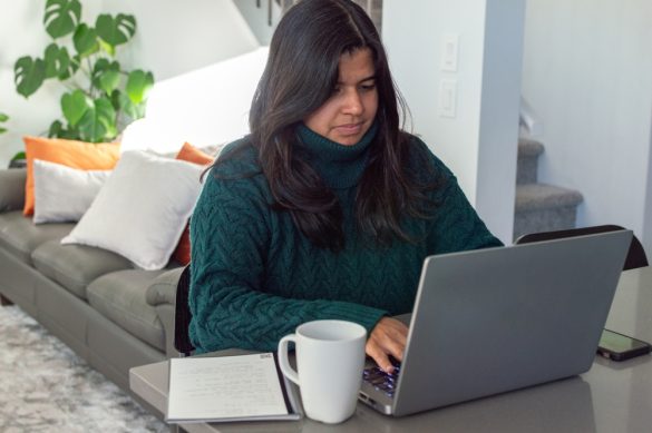 A woman wearing a jumper sits at her laptop at home with a mug next to her.