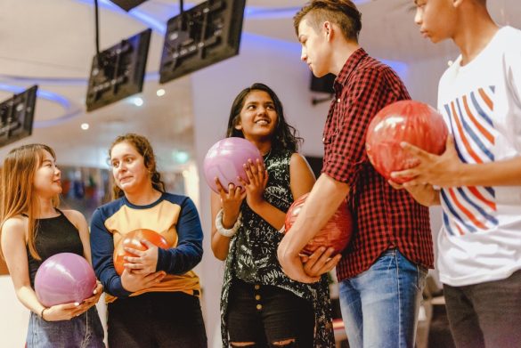 A young woman at a bowling alley holding a bowling ball, surrounded by other young people also holding bowling balls