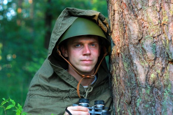 A man dressed in tactical gear and holding a pair of binoculars stands behind a tree.