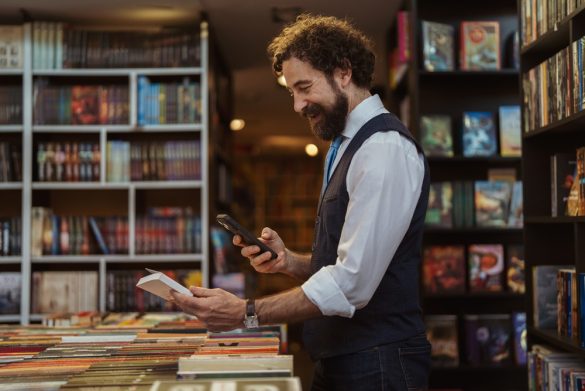 A smiling person stands in a bookshop, taking a picture of a book they are holding