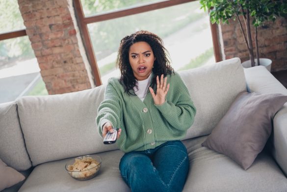 A person sitting on a sofa, pointing a TV remote towards the camera and looking frightened